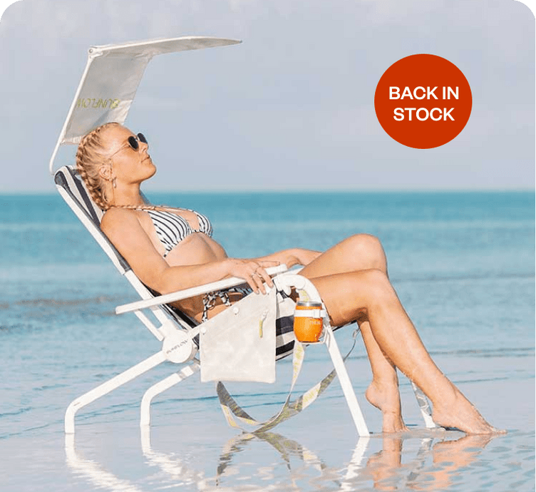 Woman relaxing on a beach chair with an umbrella and drink, ocean in the background, with a promotional label that says 'back in stock'.